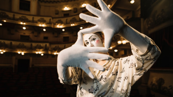 Atriz com maquiagem de mímico emoldura o palco de um grande teatro para ilustrar o Dia Mundial do Teatro 2026