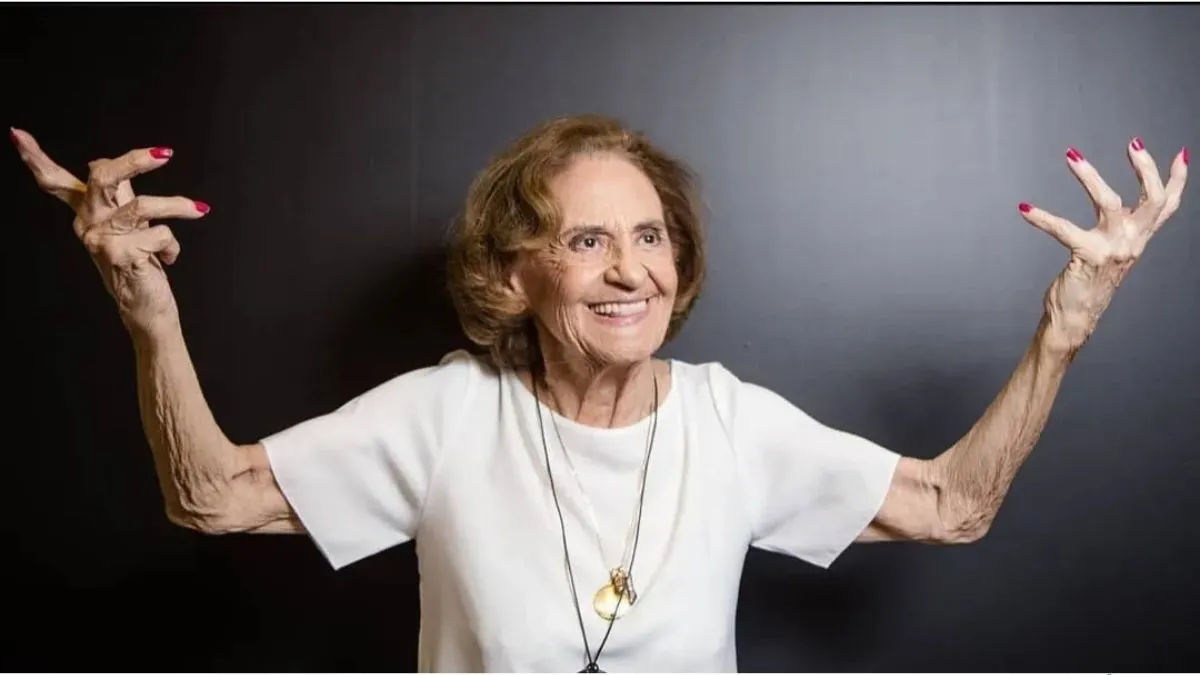 Imagem da atriz Laura Cardoso de blusa branca e de mãos levantadas com sorriso no rosto